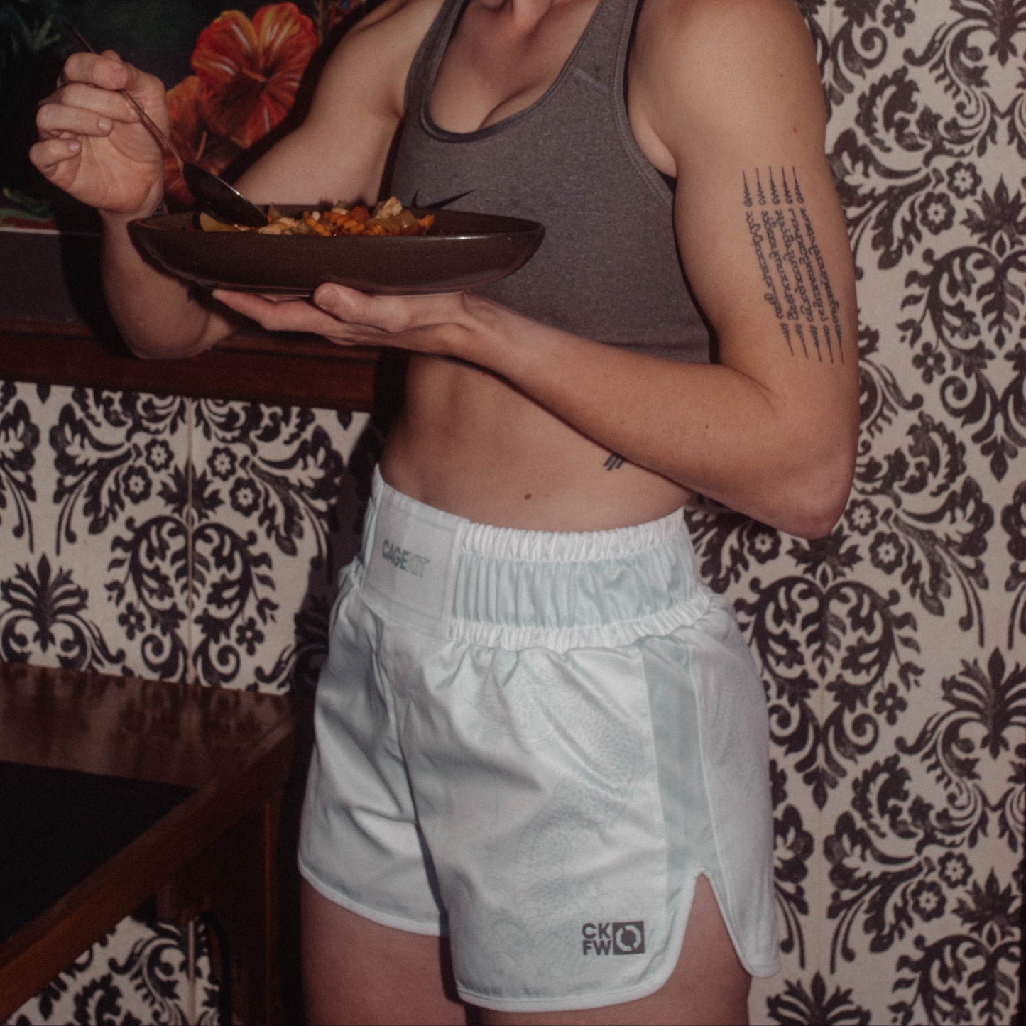 Person wearing crop top and white thai shorts with a thai tattoo on their arm, standing against a patterned background.